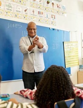 Elementary age boys and girls sitting at child-size desks and learning arithmetic from encouraging instructor in late 40s standing at board.