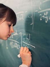 Little mathematics genius girl writing difficult mathematics equations on a green chalkboard. The gilr is smiling focused on solving the equation.