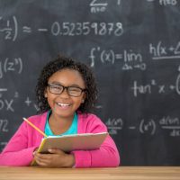 Beautiful African American elementary schoolgirl smiles confidently in classroom. Algebra equations are on the blackboard behind her.